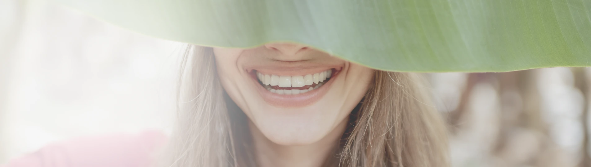 Smiling woman with clean, white teeth, partially covered by a leaf.