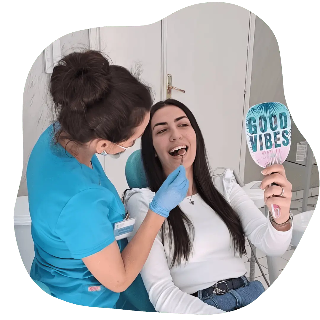 Stomatolog pruža nježan tretman dok se pacijentkinja smije i drži ogledalo sa natpisom “Good Vibes.”